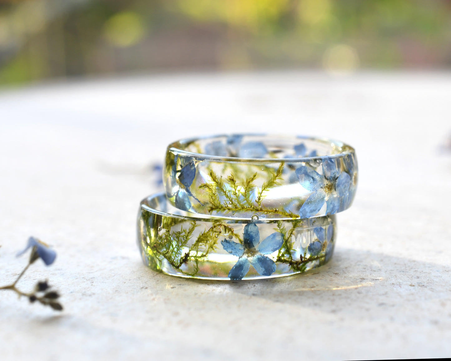 Two clear resin rings with embedded forget me not flower and dried moss on a light surface.