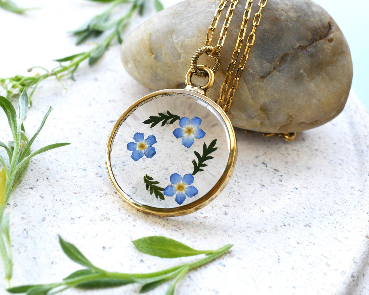 Close-up of a resin pendant with real dried forget-me-not flowers encased in clear resin, set on a gold-plated setting with a delicate chain.