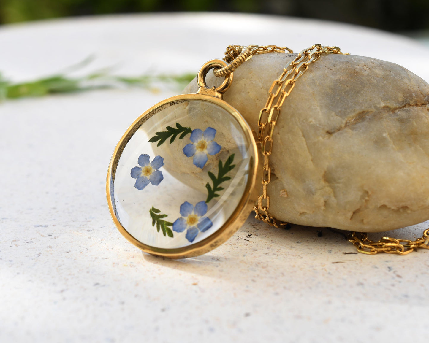 Close-up of delicate forget-me-not flowers preserved in a handmade resin pendant with a shiny gold-plated finish.