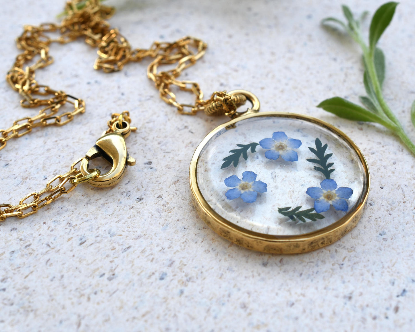 Elegant forget-me-not flower resin pendant lying on a white background, showcasing its timeless design and gold-plated chain.
