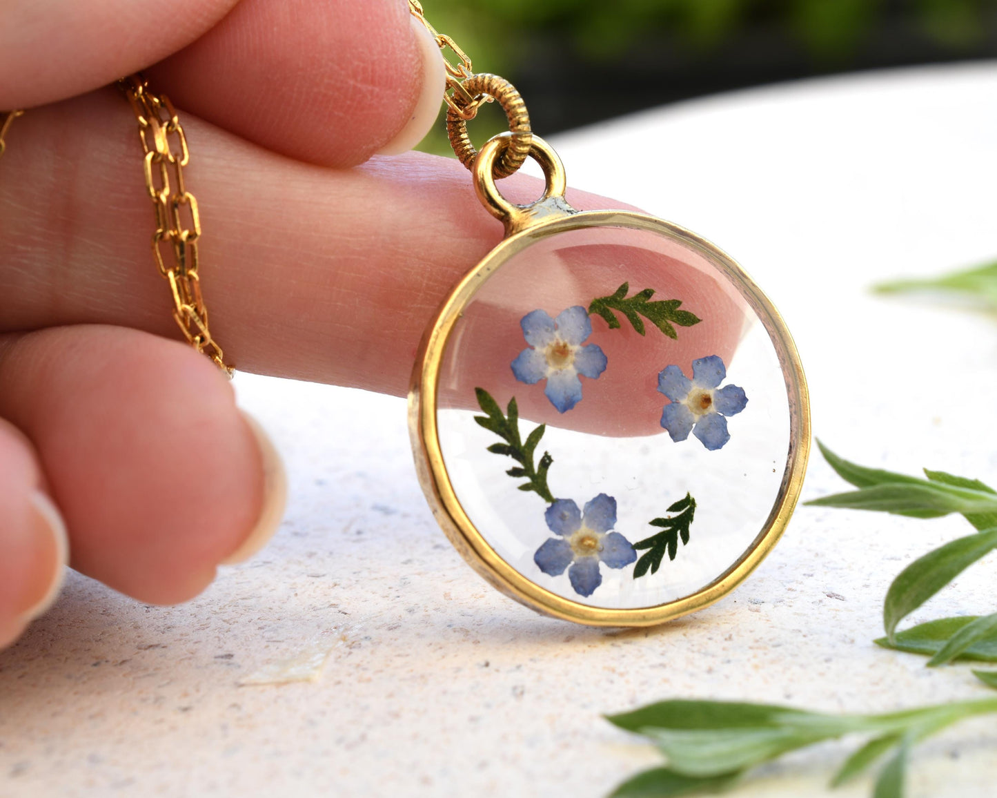 Hand holding a forget-me-not flower pendant in clear resin with a gold-plated chain, perfect for gifting or personal wear.