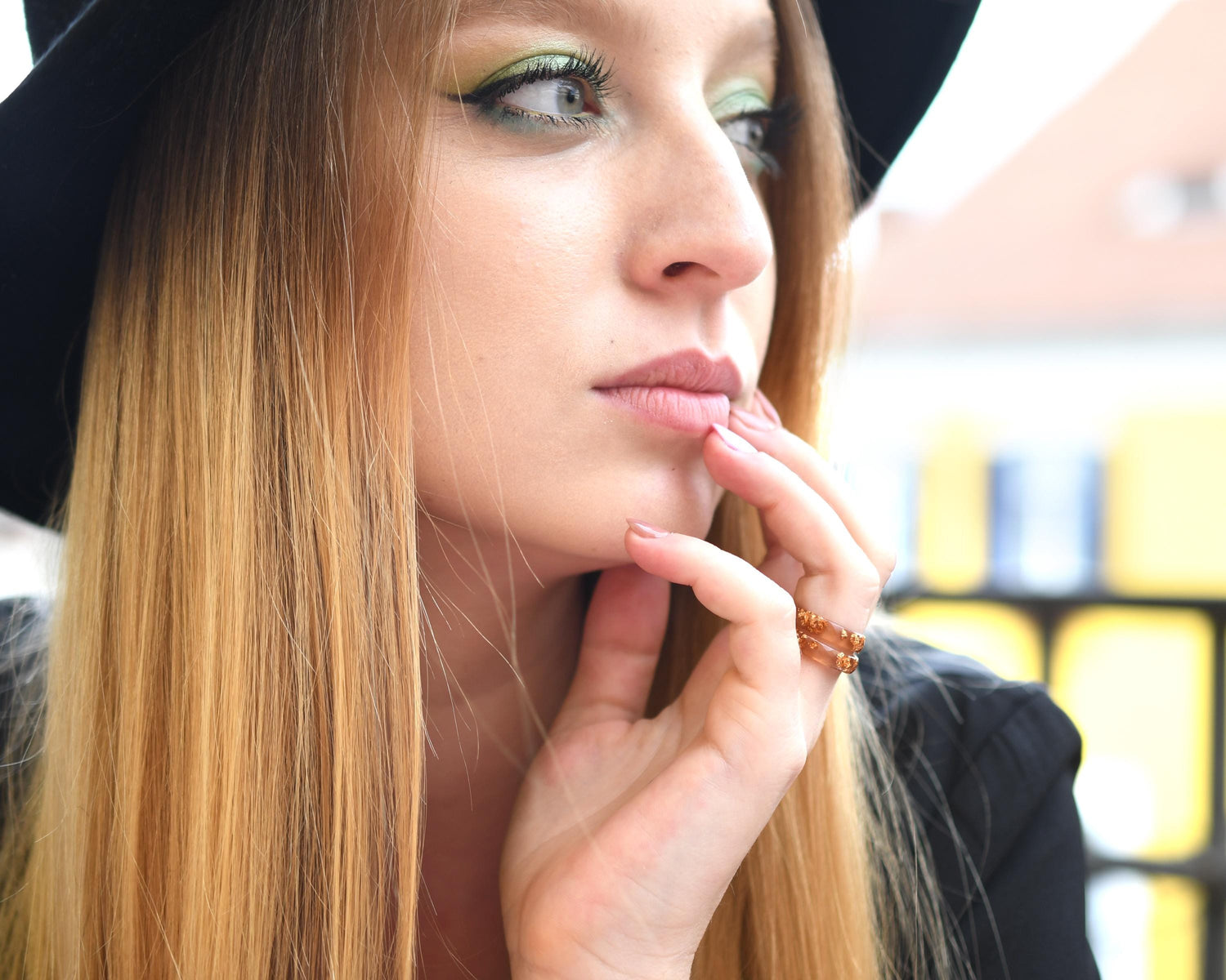 Woman with long blonde hair and a black hat, wearing a black jacket, with a blurred background.
