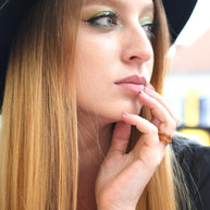 Woman with long blonde hair and a black hat, wearing a black jacket, with a blurred background.