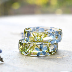 Two clear resin rings with embedded forget me not flower and dried moss on a light surface.