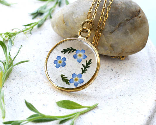 Close-up of a resin pendant with real dried forget-me-not flowers encased in clear resin, set on a gold-plated setting with a delicate chain.