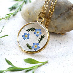 Close-up of a resin pendant with real dried forget-me-not flowers encased in clear resin, set on a gold-plated setting with a delicate chain.