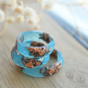 Blue spiral ring with brown accents on a wooden surface with blurred flowers in the background