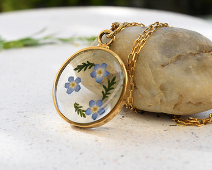 Close-up of delicate forget-me-not flowers preserved in a handmade resin pendant with a shiny gold-plated finish.