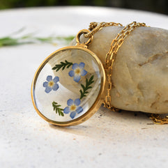 Close-up of delicate forget-me-not flowers preserved in a handmade resin pendant with a shiny gold-plated finish.