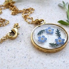 Elegant forget-me-not flower resin pendant lying on a white background, showcasing its timeless design and gold-plated chain.
