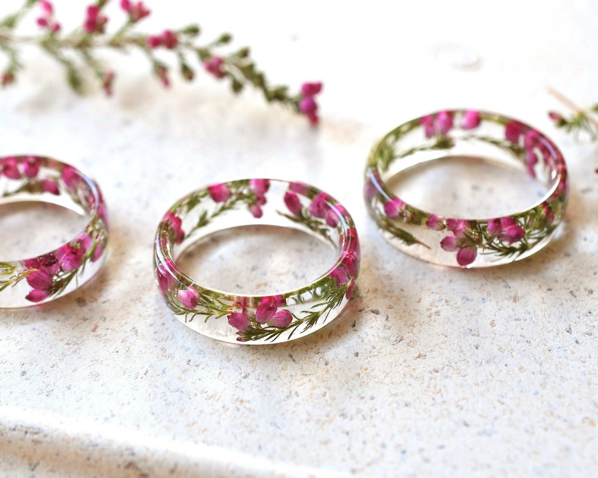 Three floral resin rings on a textured surface with pink flowers in the background.
