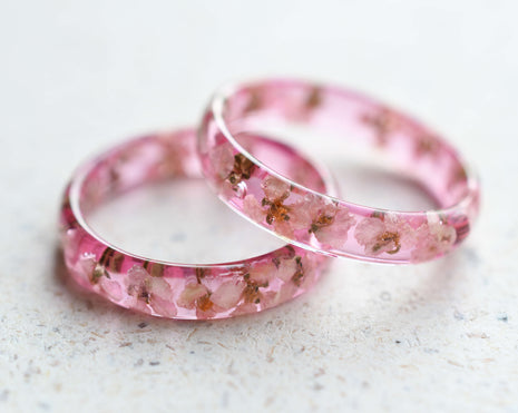 A set of two delicate pink resin rings with embedded white alyssum dried flowers, displayed on a light background.