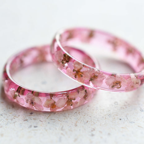A set of two delicate pink resin rings with embedded white alyssum dried flowers, displayed on a light background.