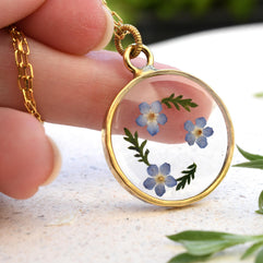 Hand holding a forget-me-not flower pendant in clear resin with a gold-plated chain, perfect for gifting or personal wear.