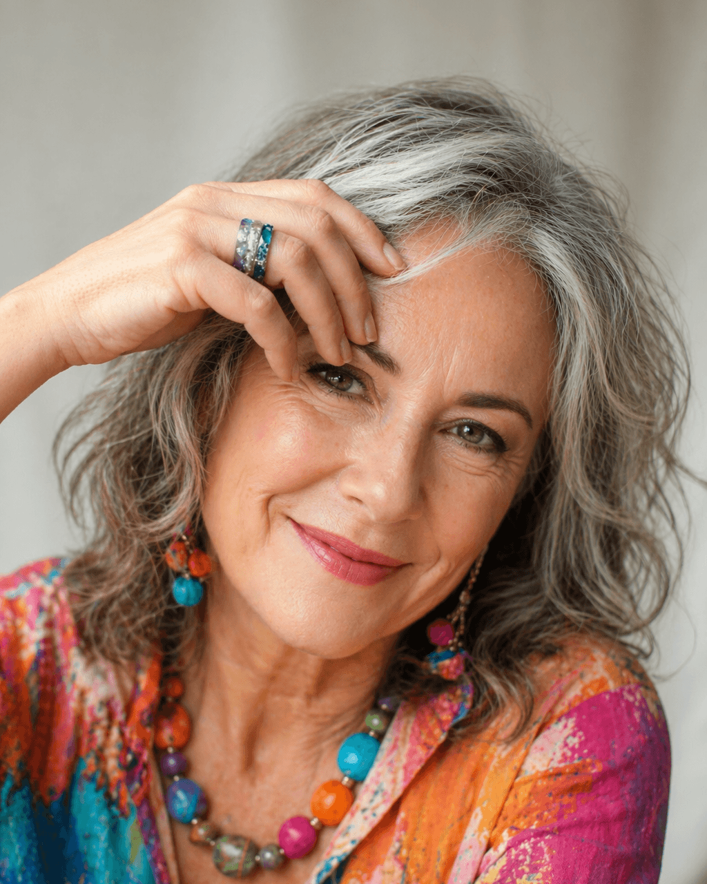 Woman with gray hair wearing colorful jewelry and a multicolored top against a neutral background