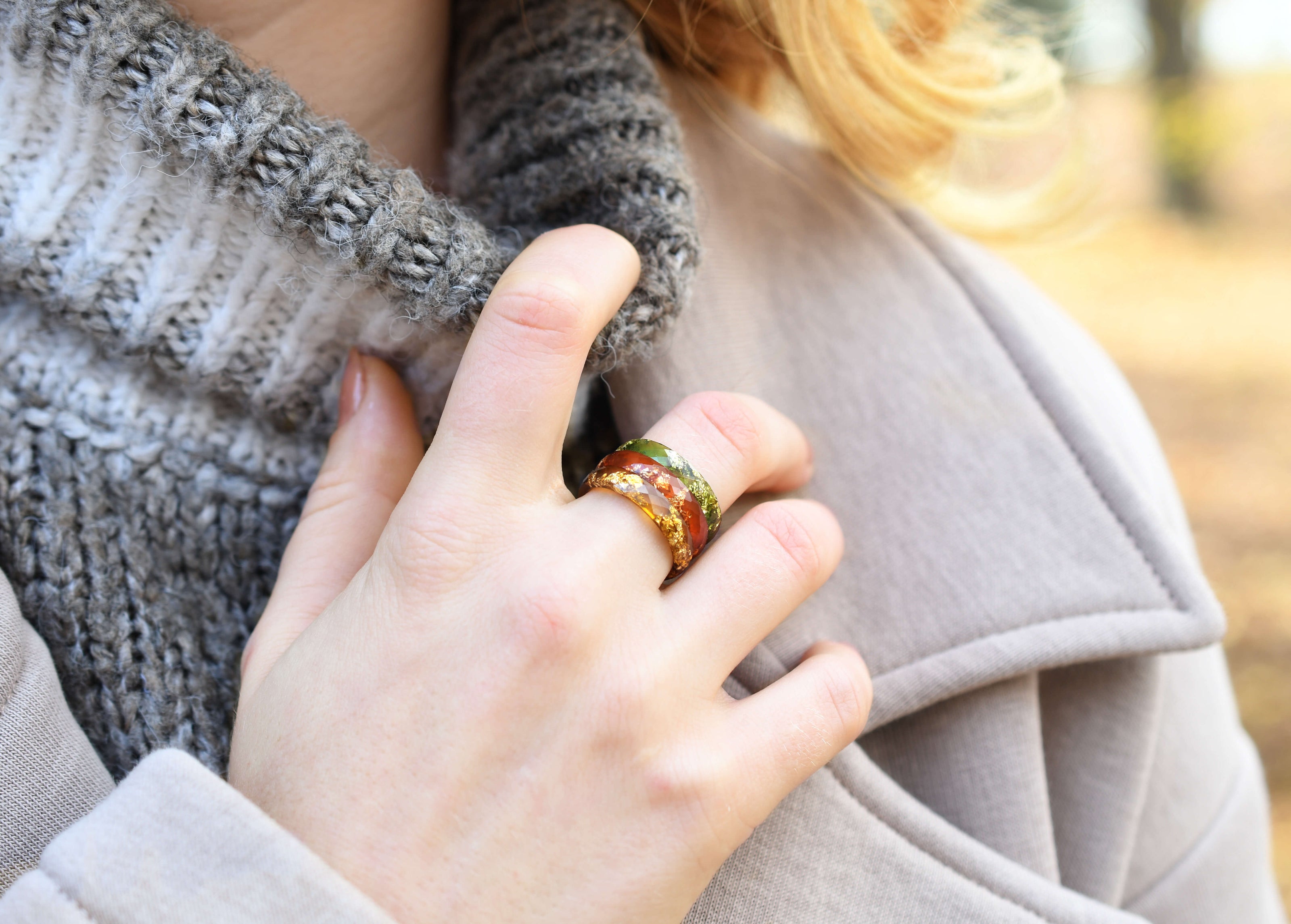 A person wearing a scarf and coat with their hand visible, showcasing three gold leaf rings on their fingers. The rings have a thin band and are set in colors that match fall hues.